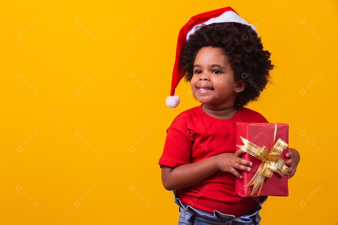 Criança afro sorridente com chapéu vermelho de Papai Noel segurando o presente de natal