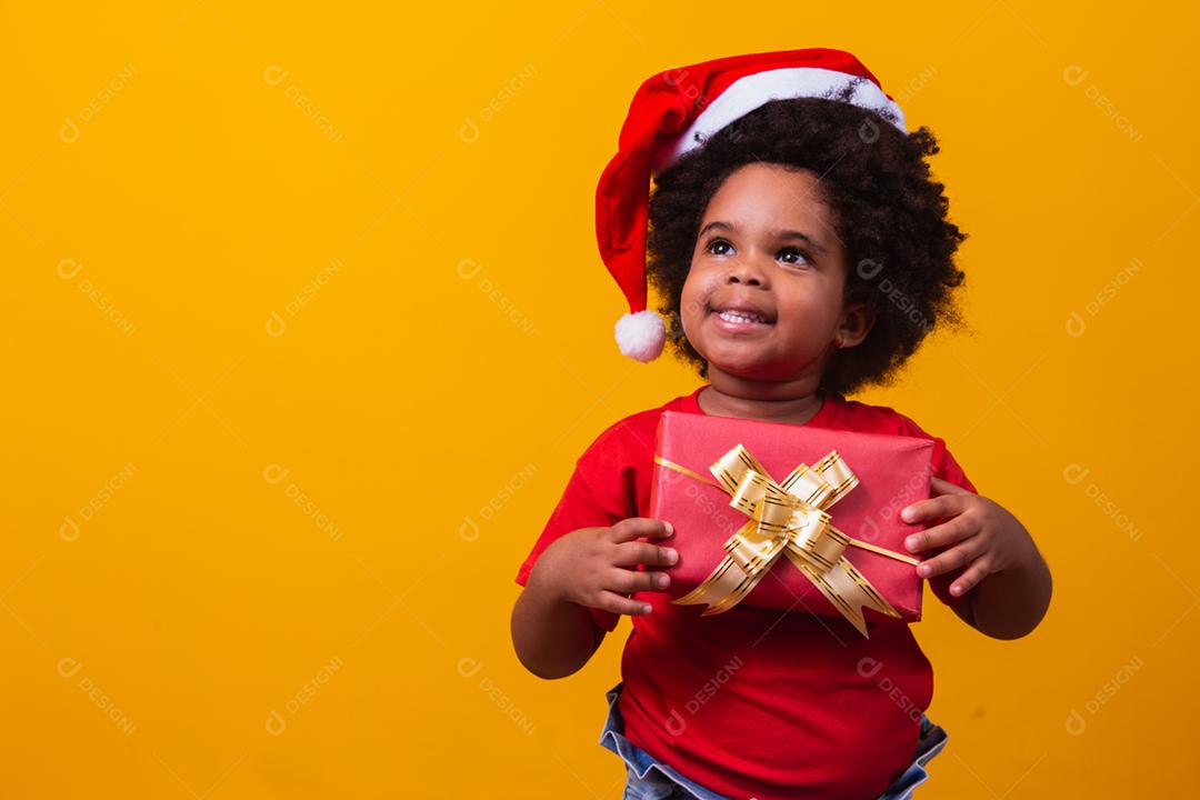 Criança afro sorridente com chapéu vermelho de Papai Noel segurando o presente de natal