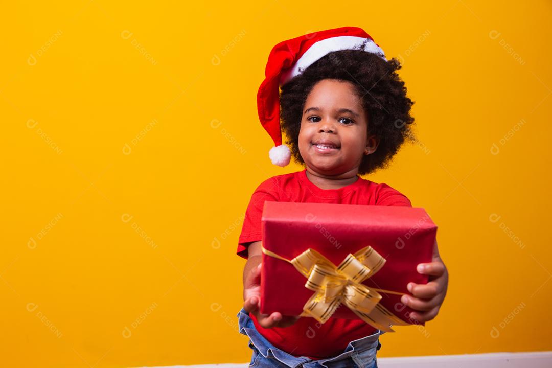 Criança afro sorridente com chapéu vermelho de Papai Noel segurando o presente de natal