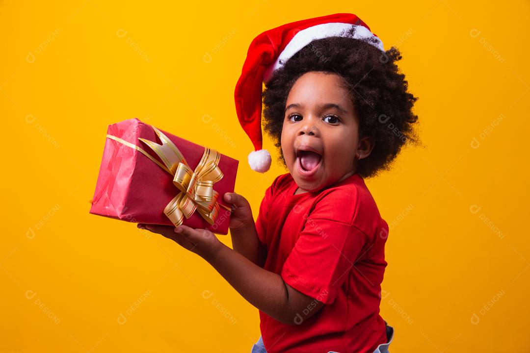 Criança afro sorridente com chapéu vermelho de Papai Noel segurando o presente de natal