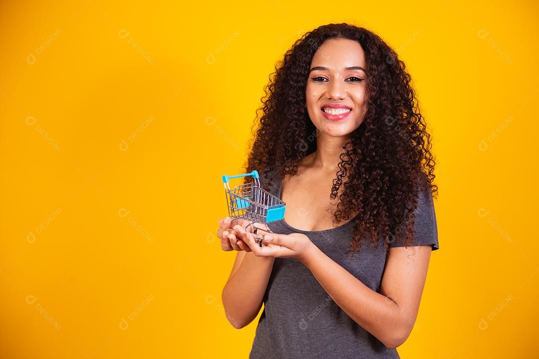 Conceito de compras. Retrato de mulher sorridente segurando pequeno carrinho de compras vazio na palma da mão, isolado em fundo amarelo