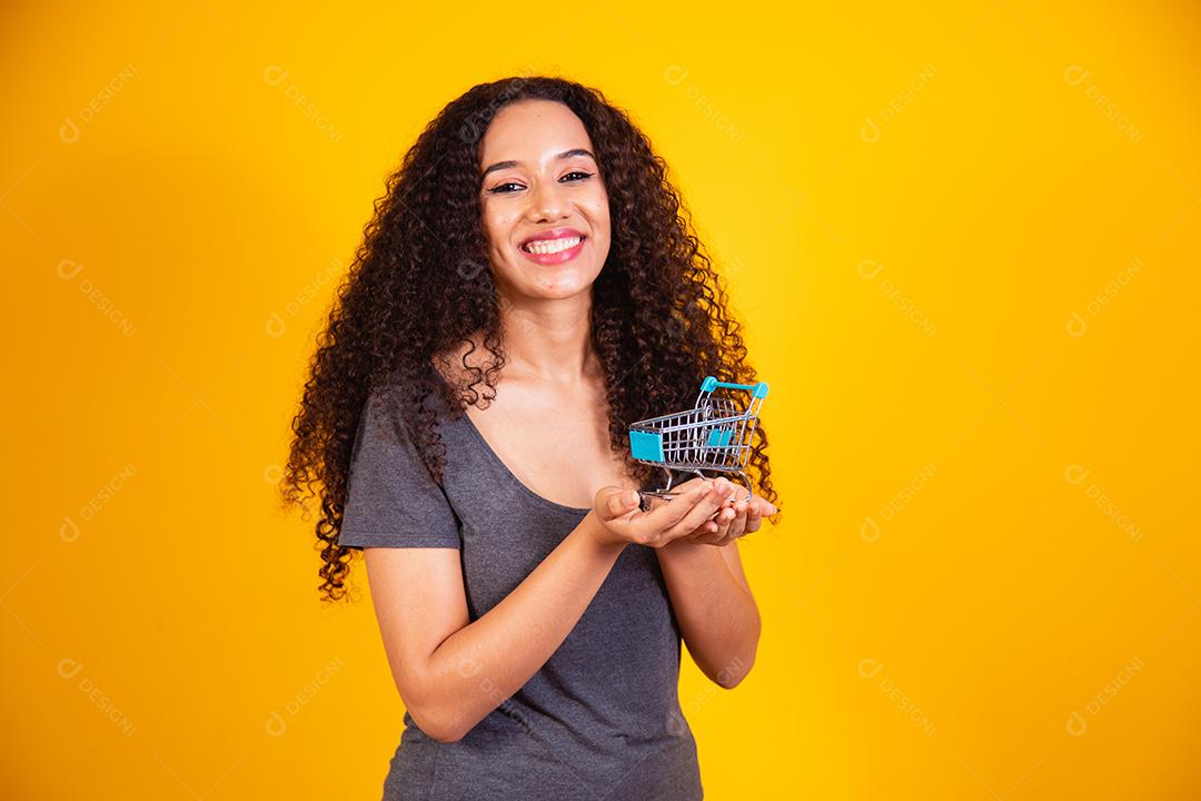 Conceito de compras. Retrato de mulher sorridente segurando pequeno carrinho de compras vazio na palma da mão, isolado em fundo amarelo