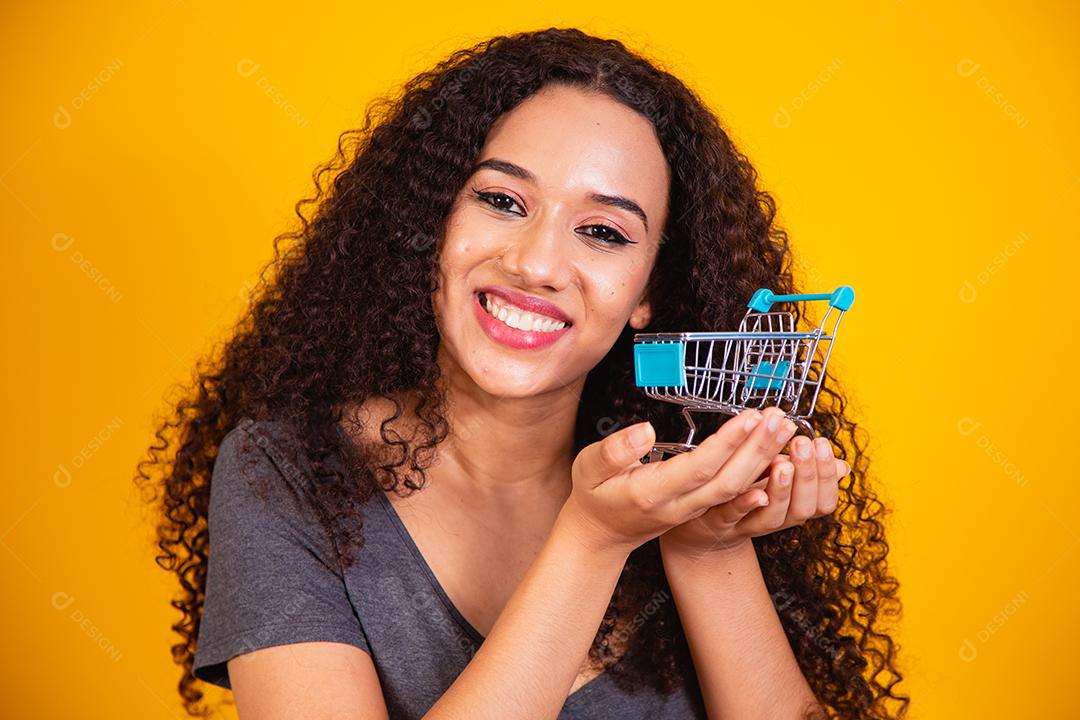 Conceito de compras. Retrato de mulher sorridente segurando pequeno carrinho de compras vazio na palma da mão, isolado em fundo amarelo