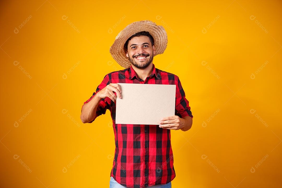 Homem brasileiro vestindo roupas típicas para a Festa Junina segurando placa