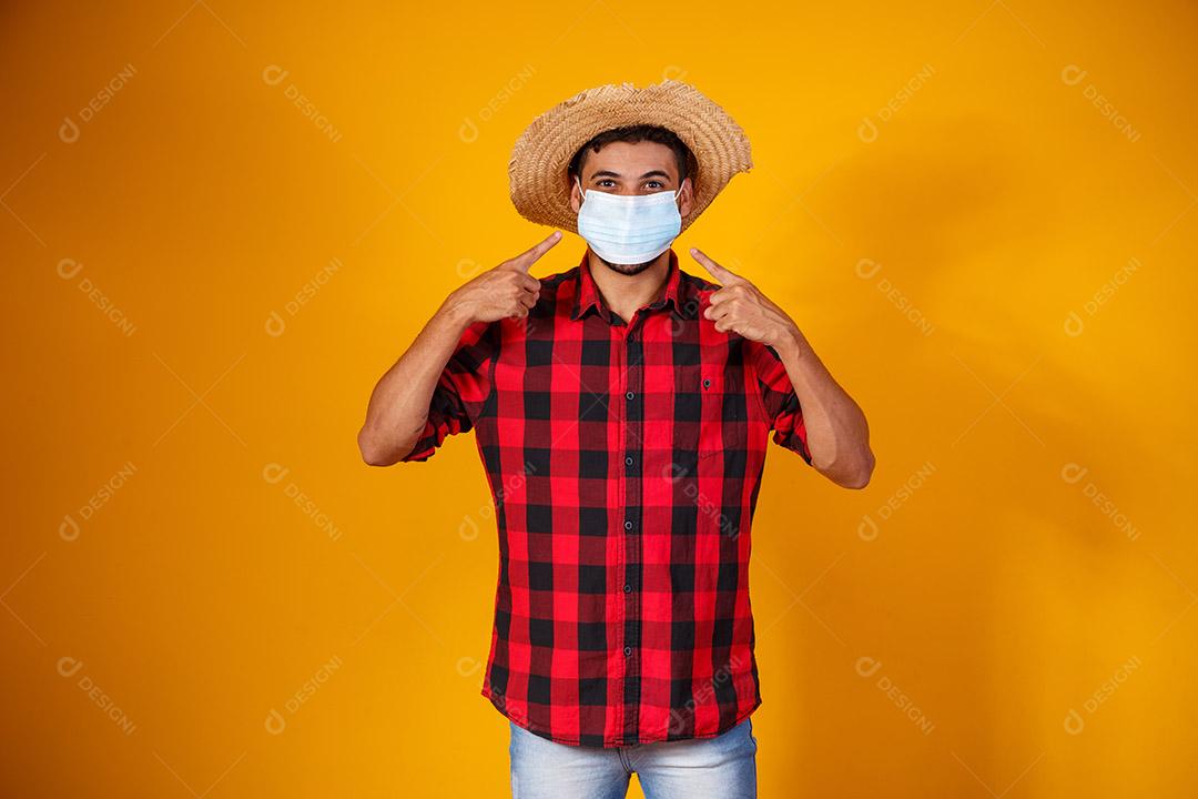 homem com roupas típicas da famosa festa brasileira chamada Festa Junina em comemoração ao São João. Menino lindo em fundo amarelo. Menino com traje de junho e máscara para prevenção do covid