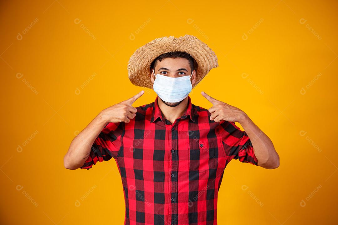 homem com roupas típicas da famosa festa brasileira chamada Festa Junina em comemoração ao São João. Menino lindo em fundo amarelo. Menino com traje de junho e máscara para prevenção do covid