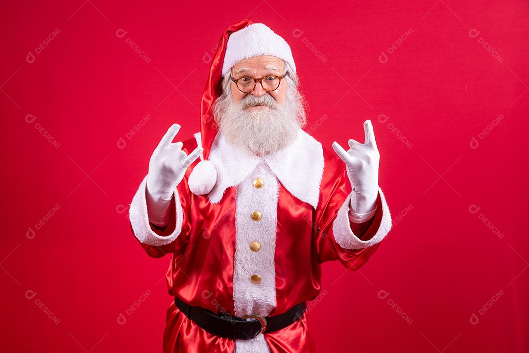 Papai Noel fazendo sinal de rock n roll com as mãos e fundo vermelho.