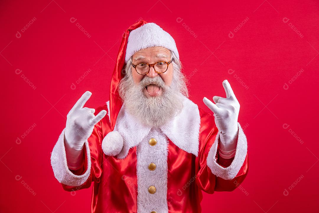 Papai Noel fazendo sinal de rock n roll com as mãos e fundo vermelho.