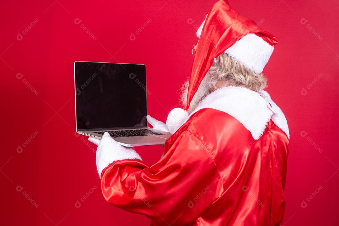 Papai Noel usando um laptop em fundo vermelho com as costas viradas.