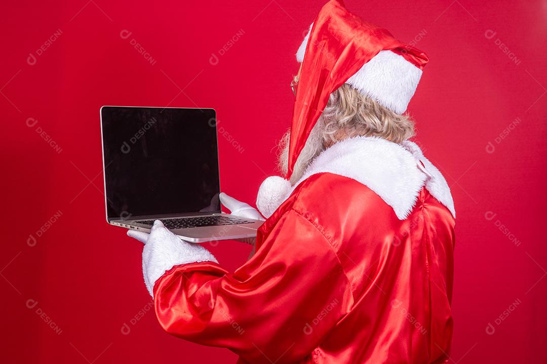 Papai Noel usando um laptop em fundo vermelho com as costas viradas.