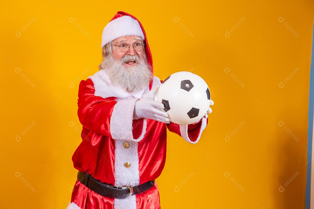 Papai Noel segurando uma bola de futebol em fundo amarelo.