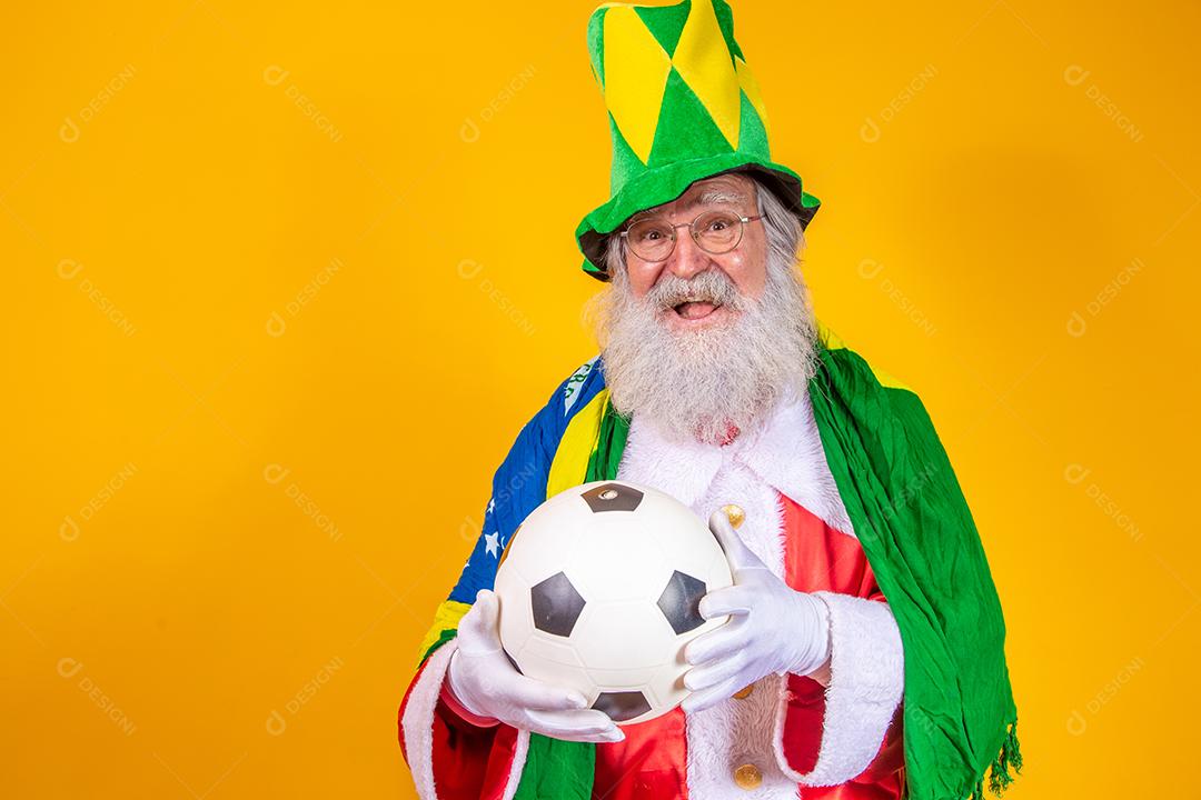 Papai Noel é fã do Brasil. Papai Noel torcedor da seleção brasileira. Campeonato esportivo. Papai Noel com a bandeira brasileira e uma bola. Partida de futebol.