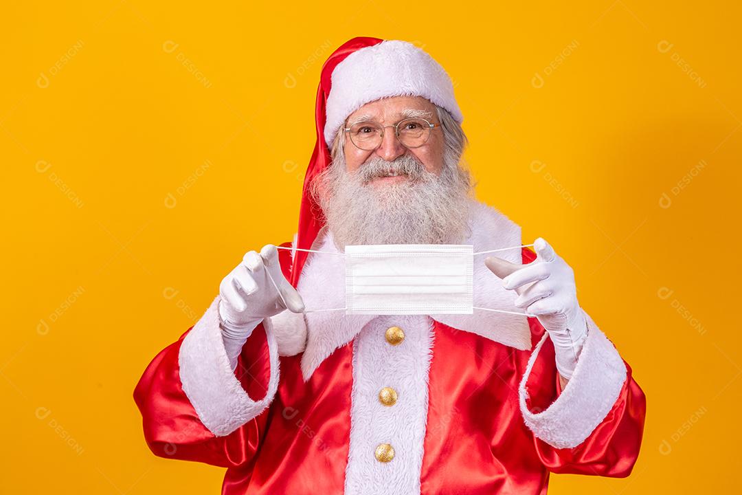 O verdadeiro Papai Noel com fundo amarelo, segurando uma máscara protetora, óculos e chapéu. Natal com distanciamento social. Covid-19