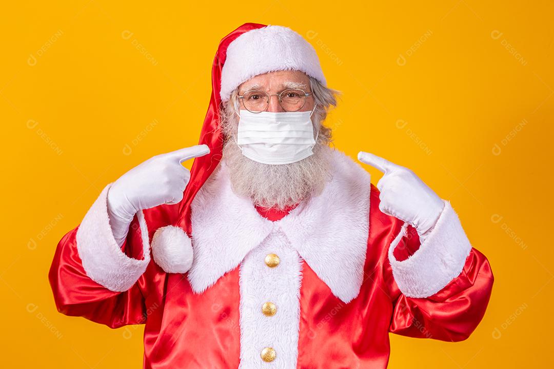 Verdadeiro Papai Noel com fundo amarelo, usando uma máscara protetora, óculos e chapéu. Natal com distanciamento social. Covid-19