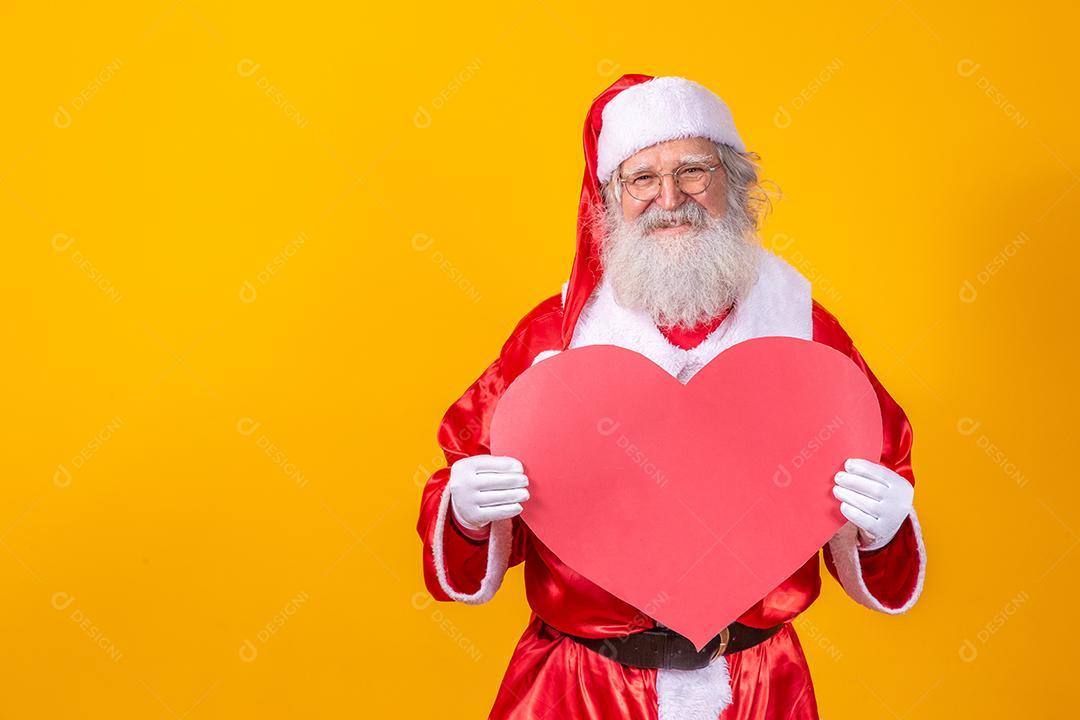 Papai Noel segurando grande cartão de coração vermelho em fundo amarelo. Papai Noel romântico.