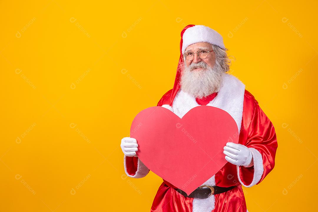 Papai Noel segurando grande cartão de coração vermelho em fundo amarelo. Papai Noel romântico.