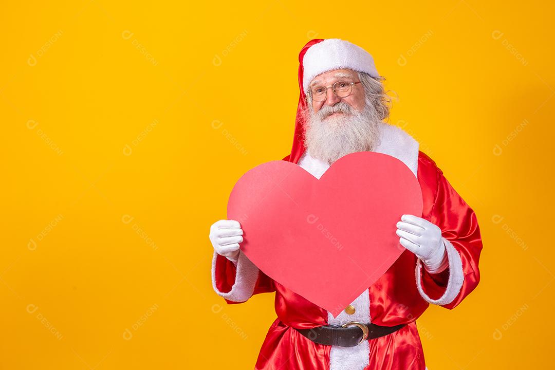 Papai Noel segurando grande cartão de coração vermelho em fundo amarelo. Papai Noel romântico.