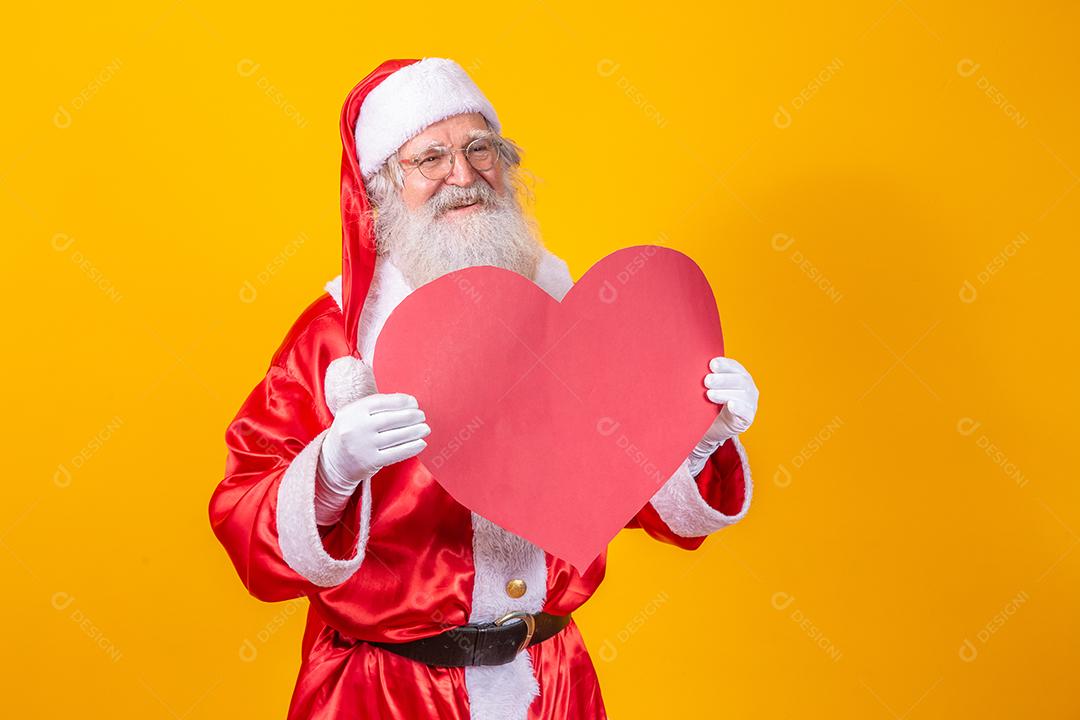 Papai Noel segurando grande cartão de coração vermelho em fundo amarelo. Papai Noel romântico.