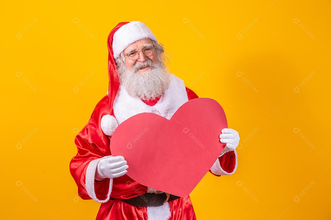 Papai Noel segurando grande cartão de coração vermelho em fundo amarelo. Papai Noel romântico.