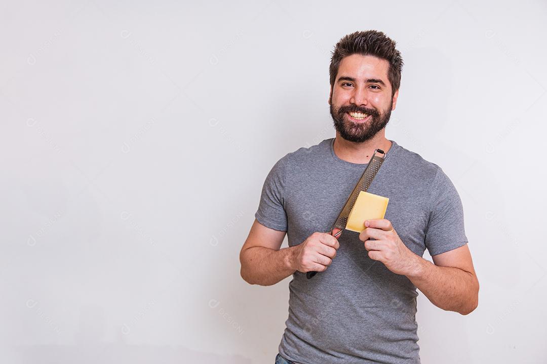 jovem segurando um ralador e um pedaço de queijo na mão sobre fundo cinza. homem vai ralar o queijo