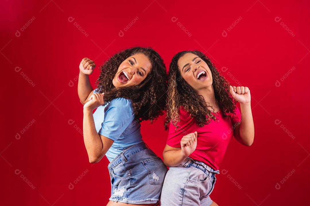 Retrato de amigos de mulheres afro jovens felizes dançando sobre fundo vermelho.