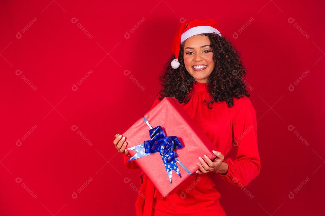 Retrato de jovem afro com chapéu de Papai Noel segurando um presente sobre fundo vermelho. conceito de noite de natal