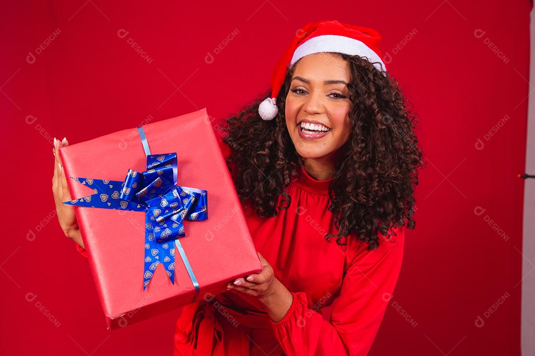 Retrato de jovem afro com chapéu de Papai Noel segurando um presente sobre fundo vermelho. conceito de noite de natal
