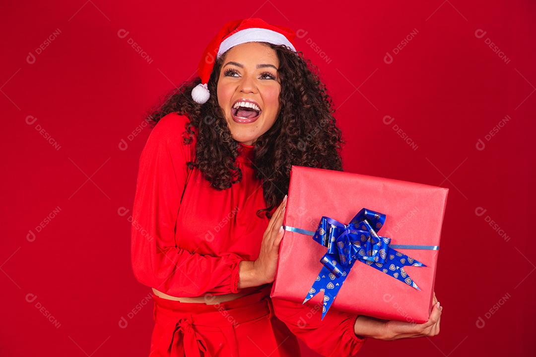 Retrato de jovem afro com chapéu de Papai Noel segurando um presente sobre fundo vermelho. conceito de noite de natal