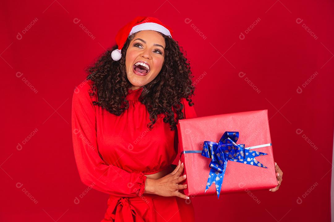 Retrato de jovem afro com chapéu de Papai Noel segurando um presente sobre fundo vermelho. conceito de noite de natal