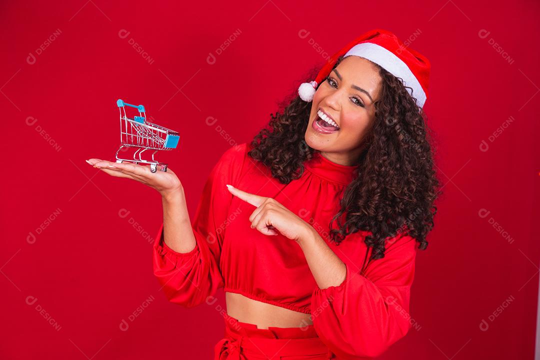 Mulher afro no chapéu de Papai Noel fazendo compras de Natal. Ela está mostrando um mini carrinho