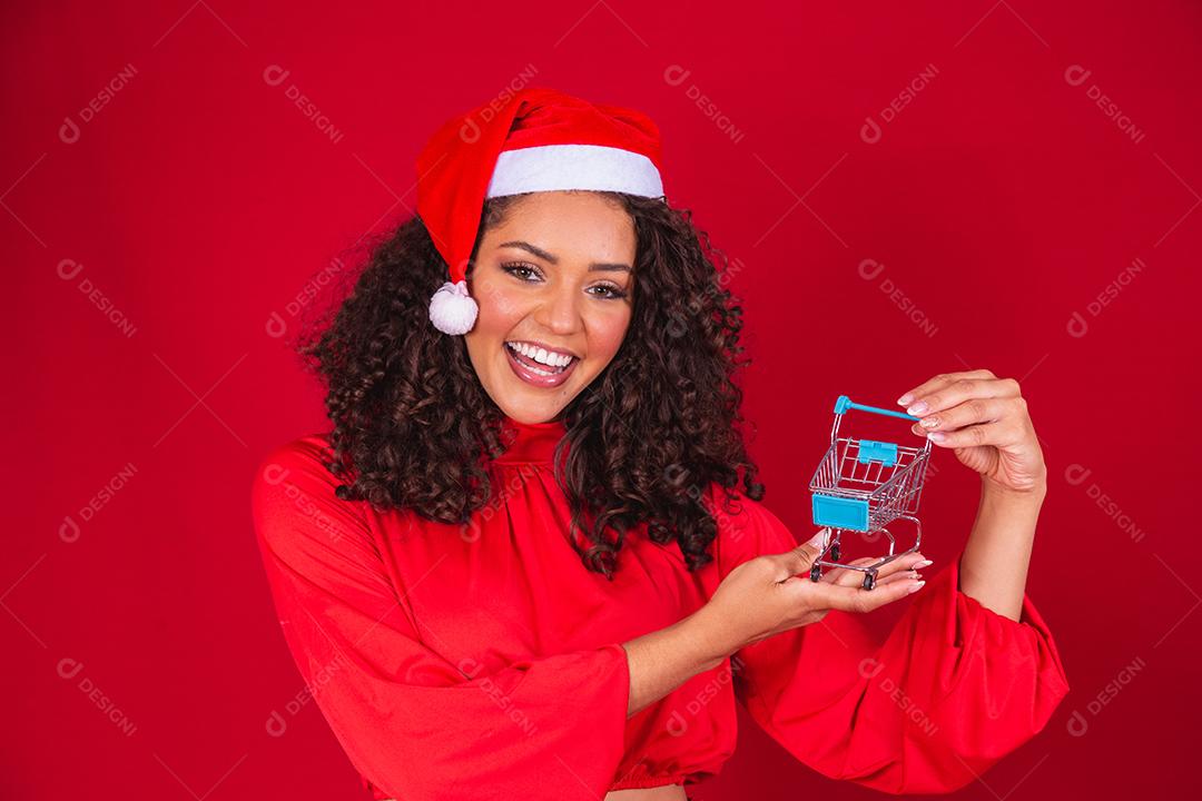 Mulher afro no chapéu de Papai Noel fazendo compras de Natal. Ela está mostrando um mini carrinho