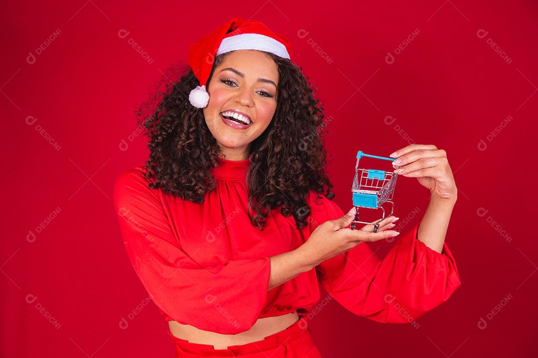 Mulher afro no chapéu de Papai Noel fazendo compras de Natal. Ela está mostrando um mini carrinho