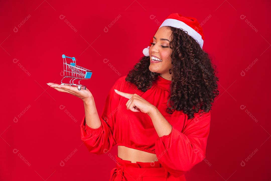 Mulher afro no chapéu de Papai Noel fazendo compras de Natal. Ela está mostrando um mini carrinho