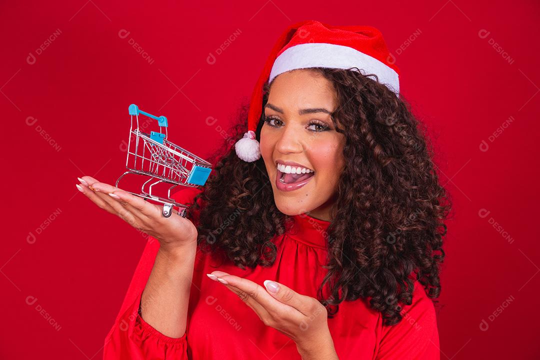 Mulher afro no chapéu de Papai Noel fazendo compras de Natal. Ela está mostrando um mini carrinho