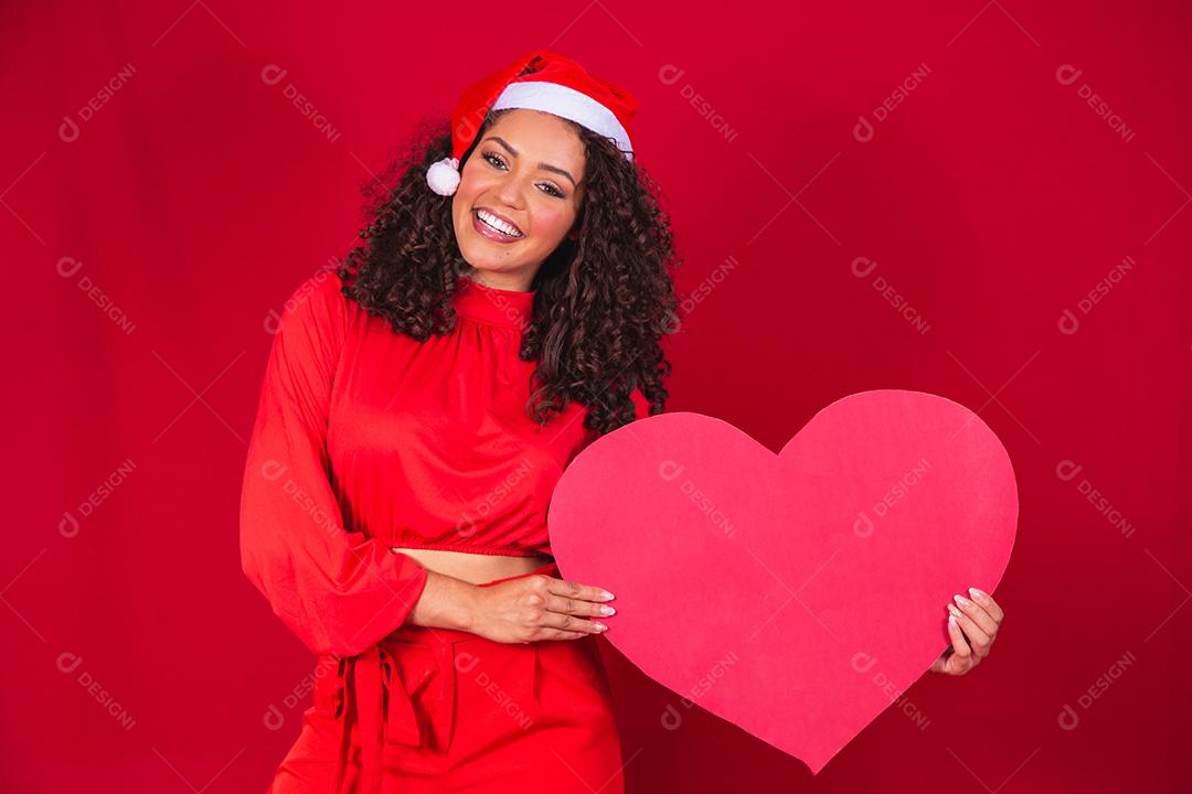 Sorridente jovem afro com chapéu de natal segurando um cartão de coração enorme