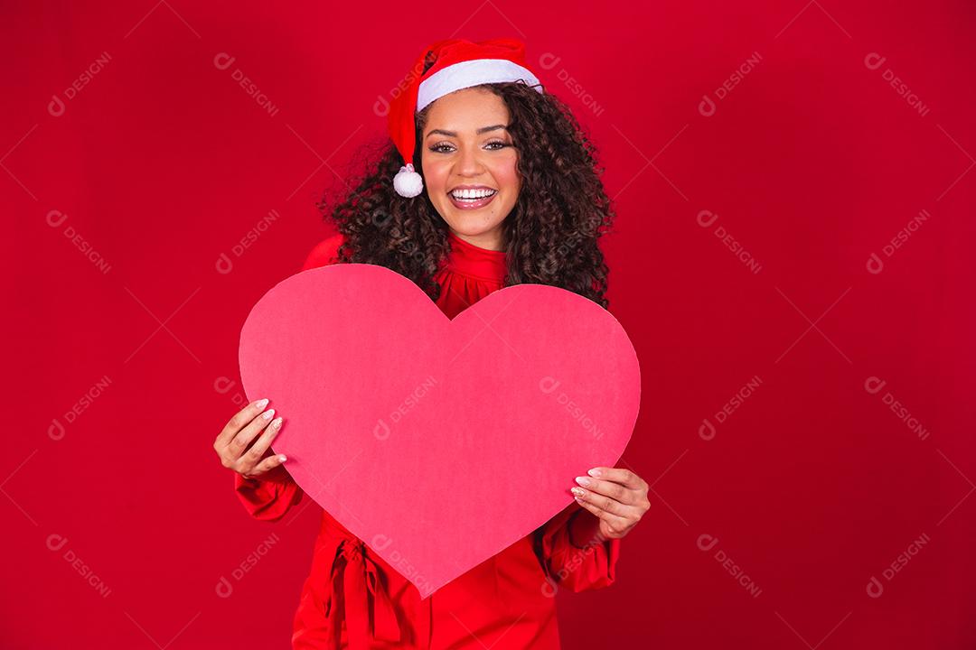 Sorridente jovem afro com chapéu de natal segurando um cartão de coração enorme