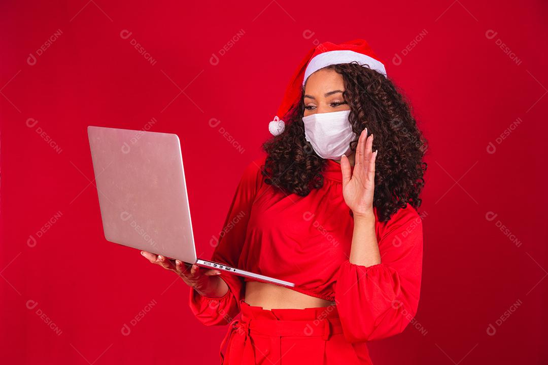 Mulher de Máscara comemorando o natal em fundo vermelho e fazendo uma chamada de vídeo no laptop