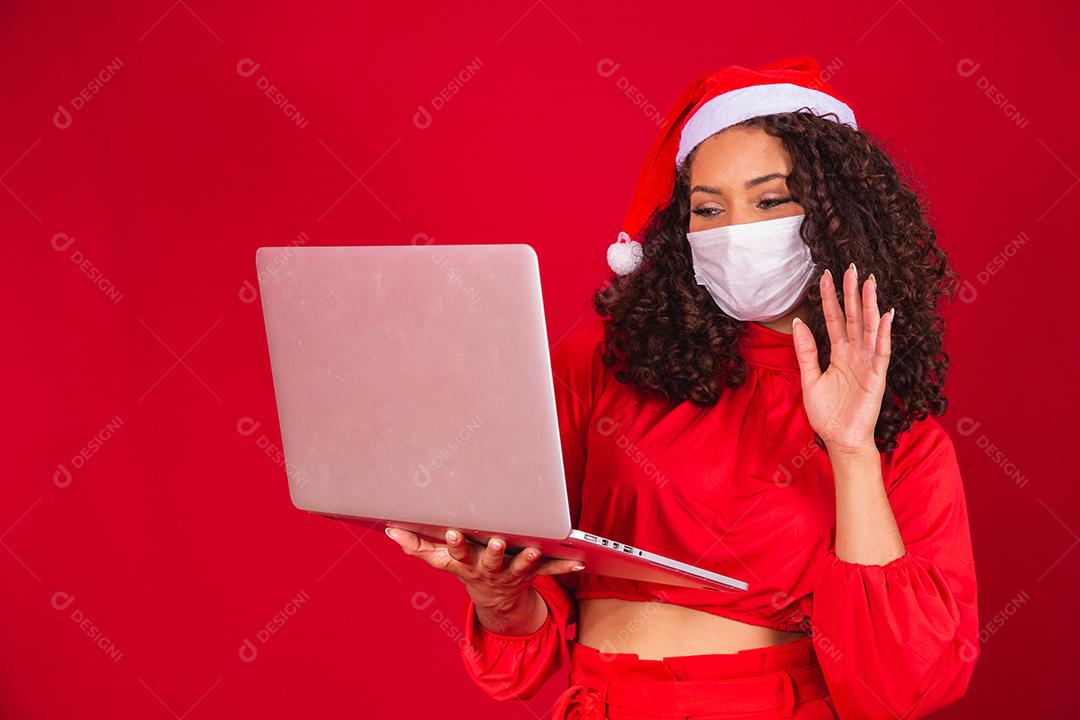 Mulher de Máscara comemorando o natal em fundo vermelho e fazendo uma chamada de vídeo no laptop