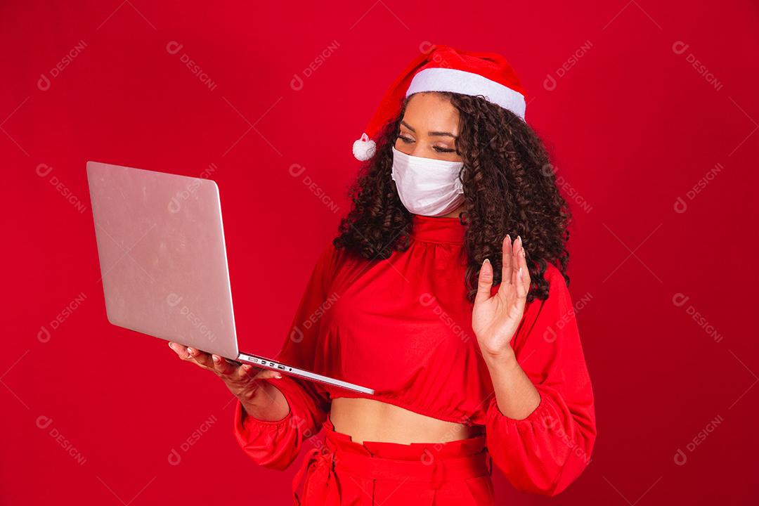 Mulher de Máscara comemorando o natal em fundo vermelho e fazendo uma chamada de vídeo no laptop