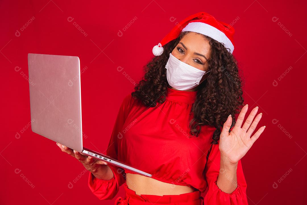 Mulher de Máscara comemorando o natal em fundo vermelho e fazendo uma chamada de vídeo no laptop