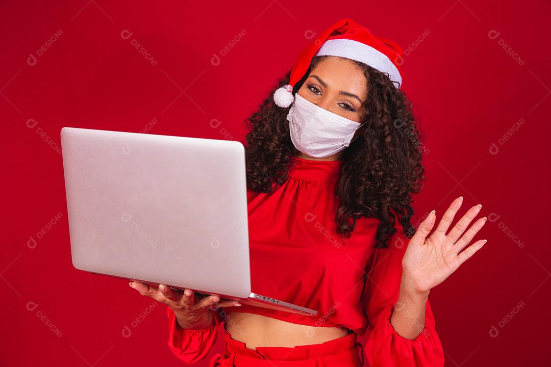 Mulher de Máscara comemorando o natal em fundo vermelho e fazendo uma chamada de vídeo no laptop