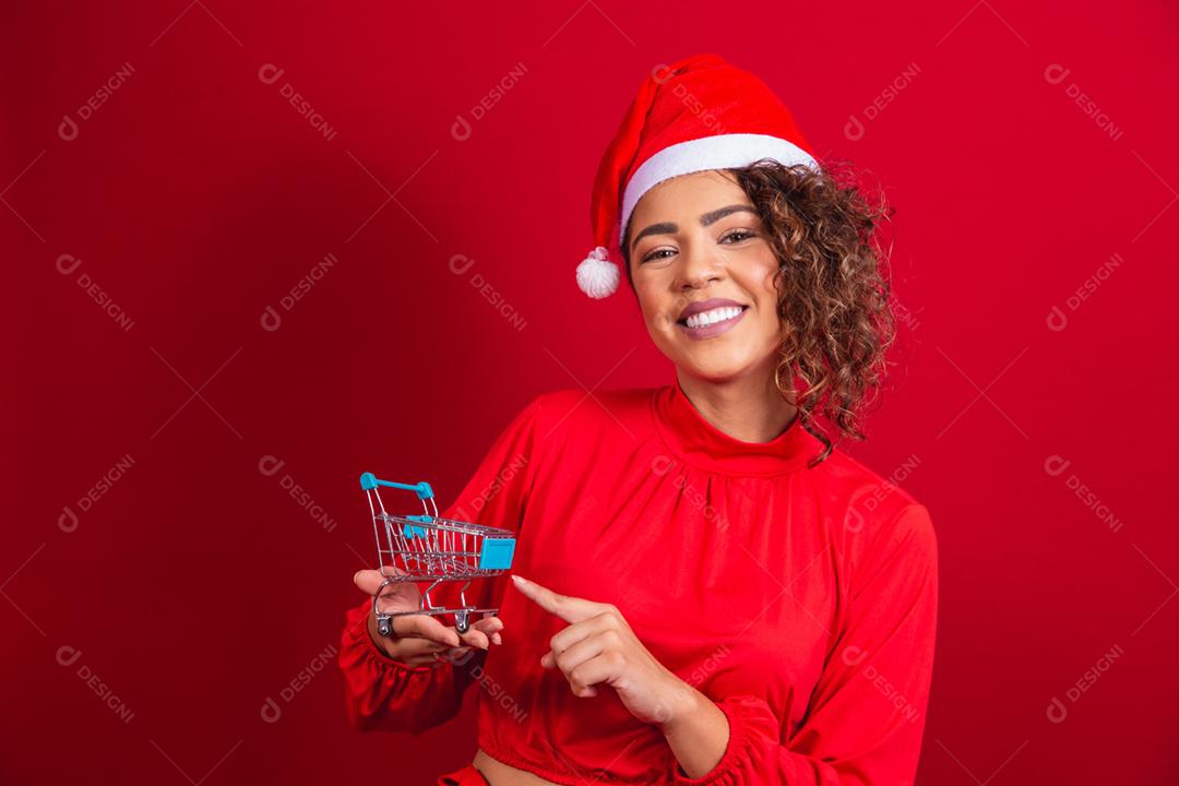 Mulher afro no chapéu de Papai Noel fazendo compras de Natal. Ela está mostrando