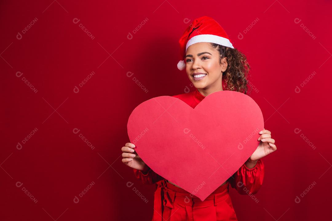 Sorridente jovem afro com chapéu de natal segurando um coração enorme