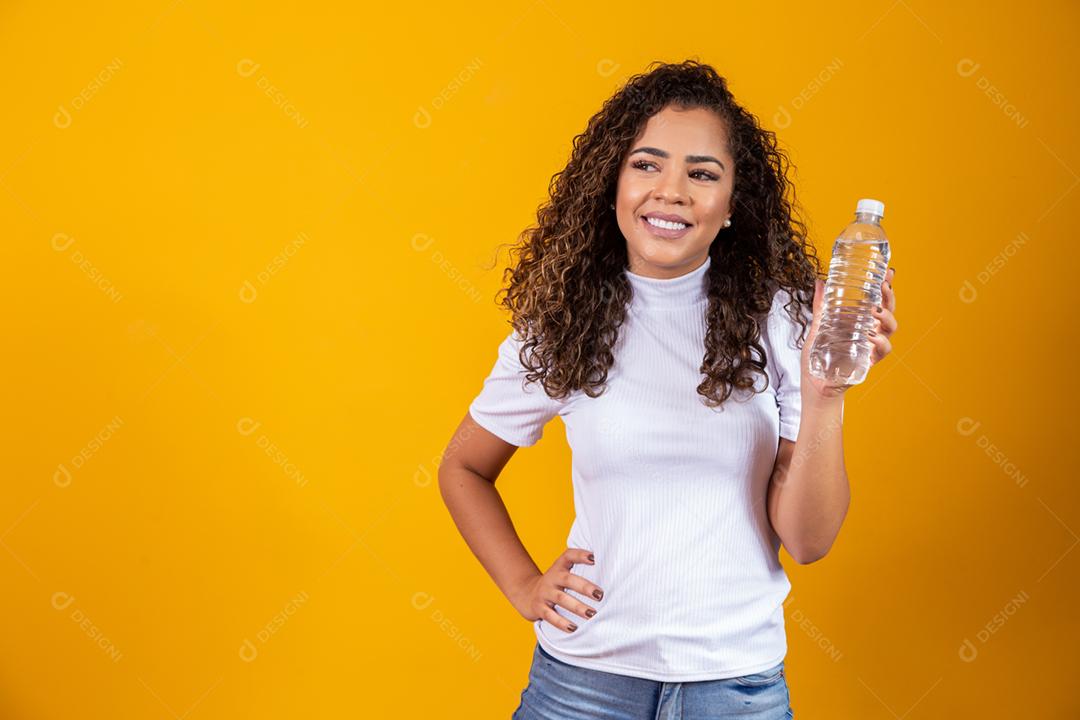 Mulher segurando uma garrafa de água em fundo amarelo. Beber água