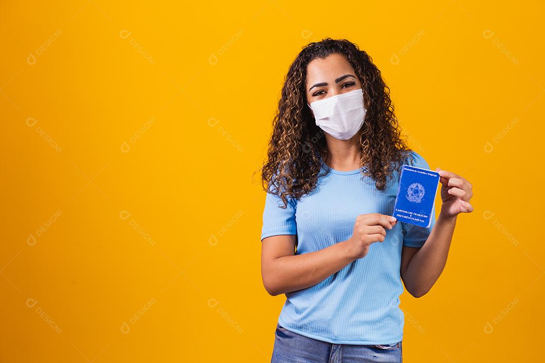 mulher afro com máscara cirúrgica segurando o cartão de trabalho brasileiro em fundo amarelo. Trabalho, economia e conceito de pandemia