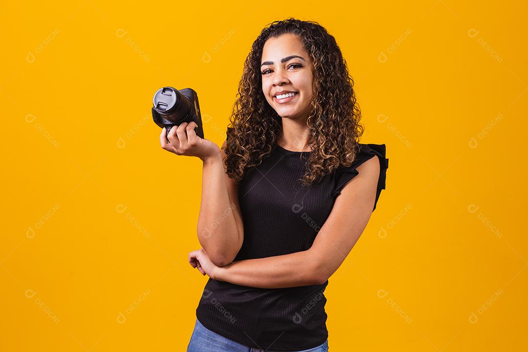 Fotografia jovem afro em fundo amarelo, segurando uma câmera fotográfica.