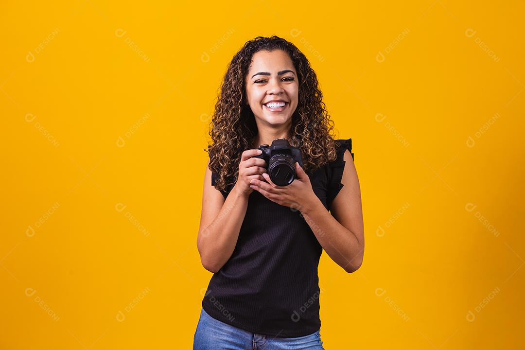 Fotografia jovem afro em fundo amarelo, segurando uma câmera fotográfica.