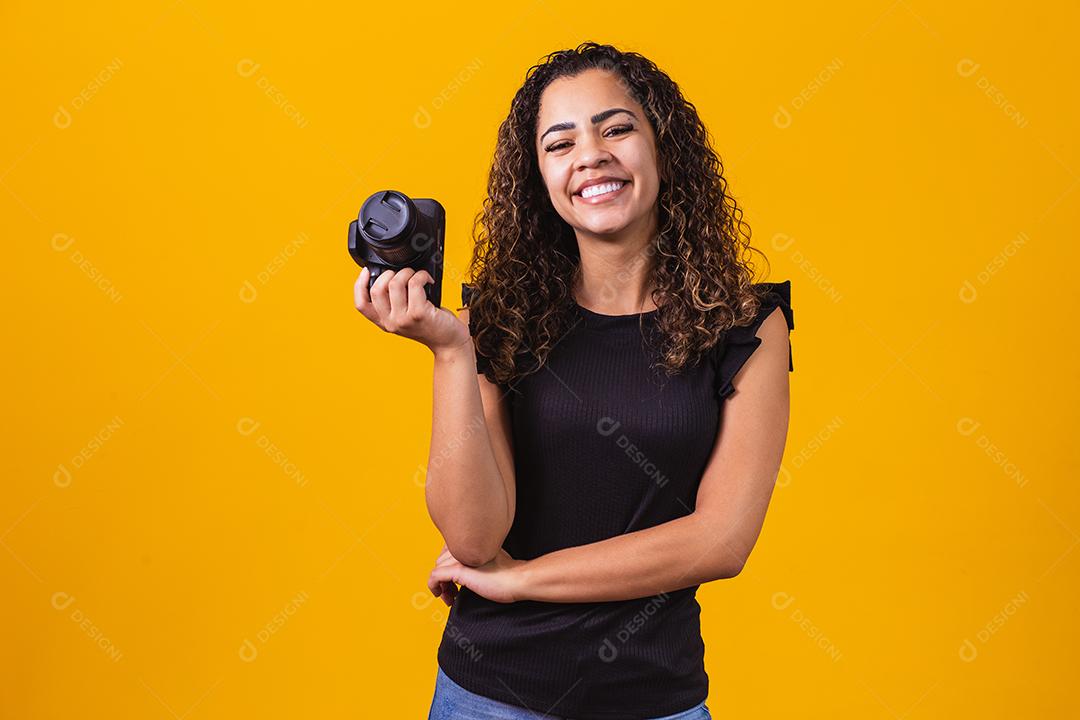 Fotografia jovem afro em fundo amarelo, segurando uma câmera fotográfica.