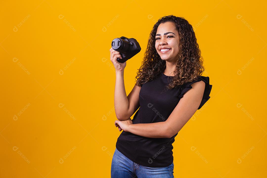 Fotografia jovem afro em fundo amarelo, segurando uma câmera fotográfica.