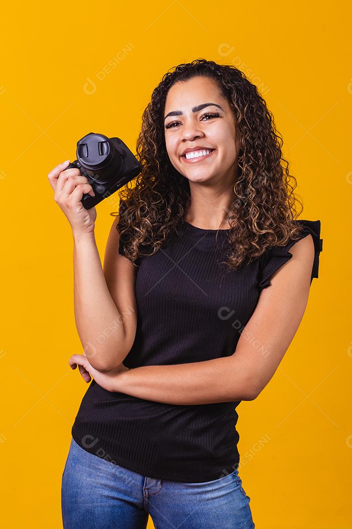 Fotografia jovem afro em fundo amarelo, segurando uma câmera fotográfica.